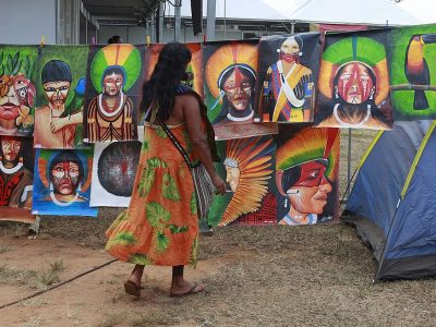 Woman in a patterned orange dress looks at a display of colourful paintings depicting Indigenous people, traditional dress and tropical birds at an outdoor art market.