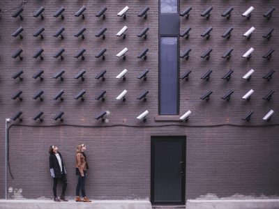 Women observing a wall covered with numerous security cameras in an urban setting.