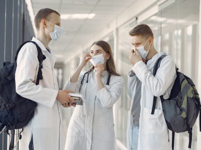 Three young medical professionals discuss in a hospital corridor wearing protective masks.