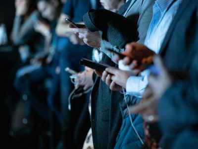 Several people in business attire stand close together while looking at and using smartphones. The image focuses on their hands and devices, with the background softly blurred.