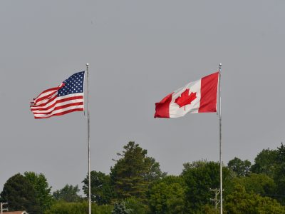 Flags of the USA and Canada waving on flagpoles against a clear grey sky