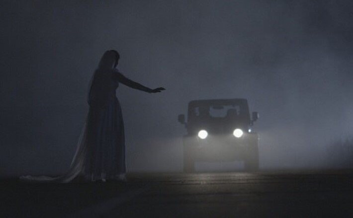 A woman hitch hiker dressed in a wedding dress asking an approach car for assistance at night.