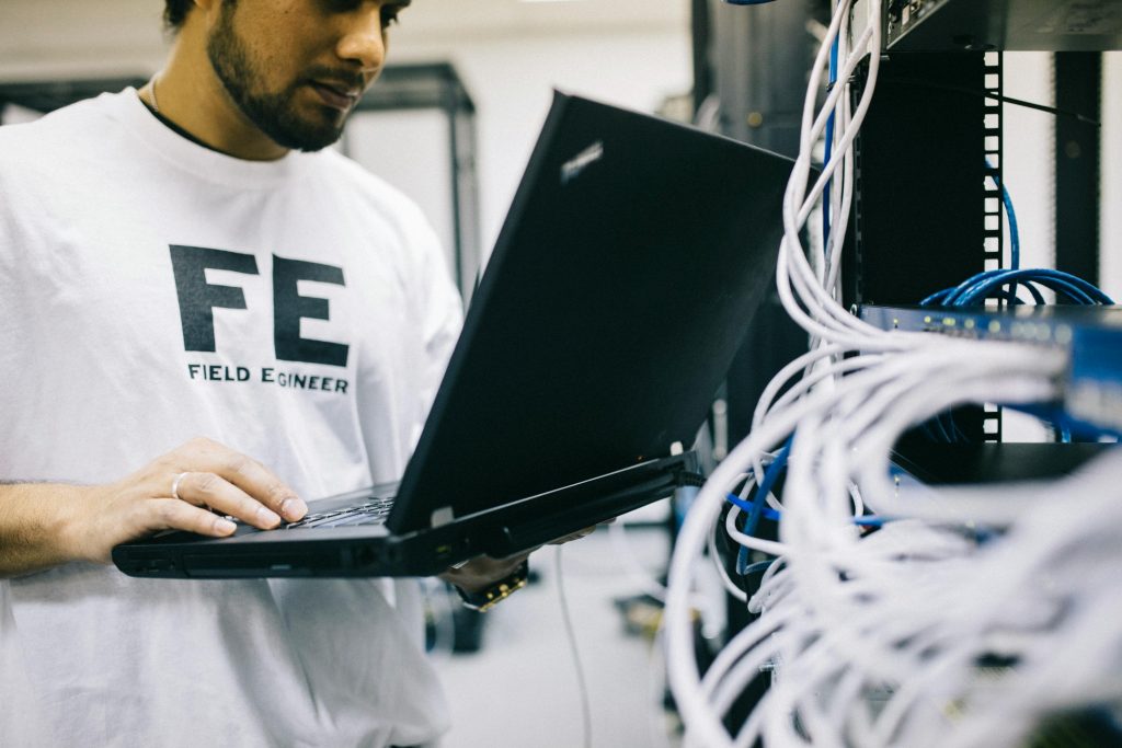 A man wearing a T-shirt that says "field engineer," while he is working on a laptop. 
