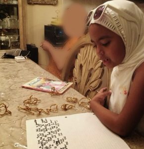 Little girl wearing a white hijab, reading a story she wrote on a piece of white lined paper.