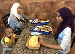 A young girl with a purple hijab, a mother with a white hijab and a baby boy are sitting eating their dinner at a restaurant.