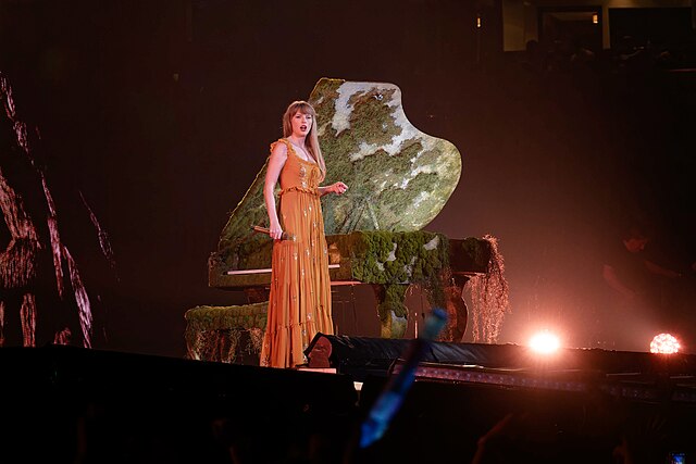 Photo of Taylor Swift on stage next to a moss-covered piano.