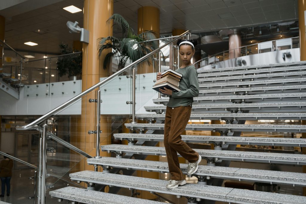 A young girl walking down the stairs of a library with a stack of books in her hand