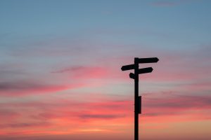 A sign with different directions and a sunset in the back.